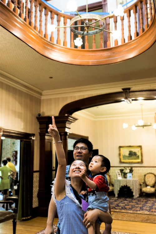 A young couple with the lady holding a toddler while pointing up with her hand with everyone looking up in the main hall of the museum in Government House SK