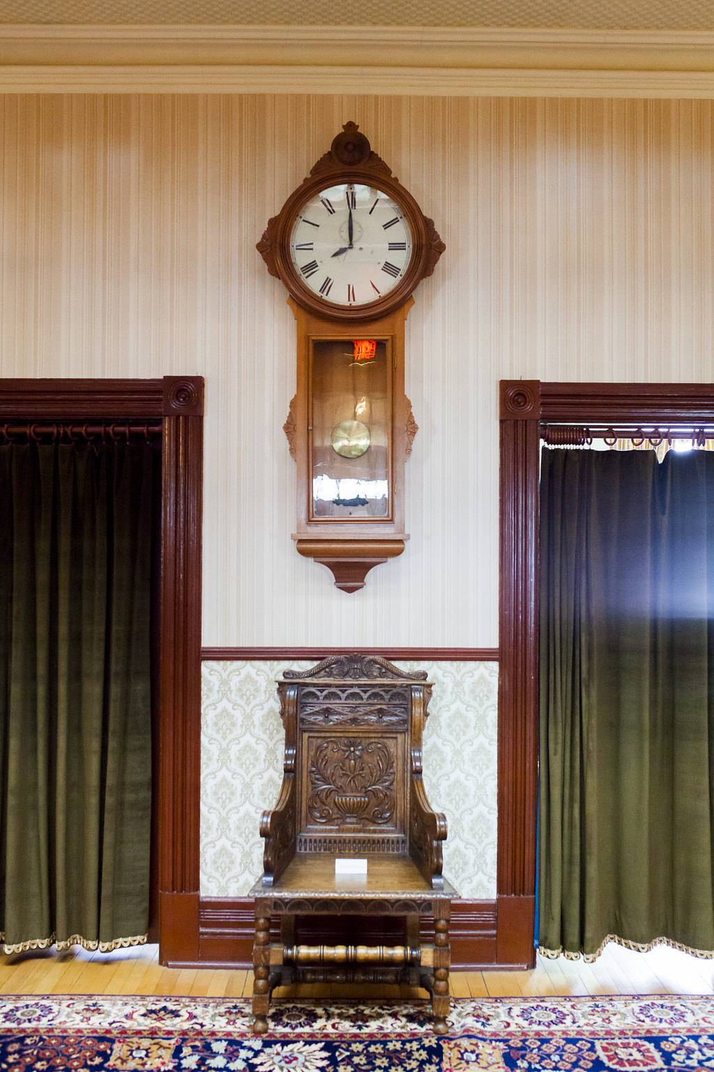 A wooden clock that is original to Government House SK hangs on a wall above a wooden chair in the main hall of the museum