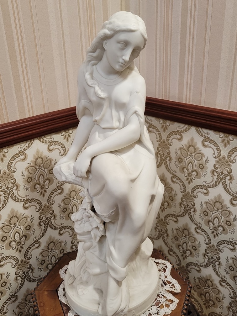 A white statue of a woman sitting on a log which sits on a table in the museum of Government House SK
