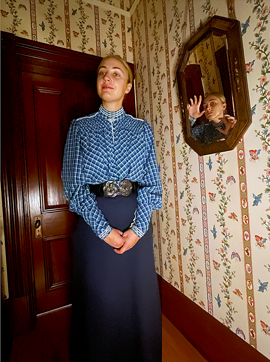 A young woman dressed in Victorian costume standing in front of a mirror that has a scary image of her.