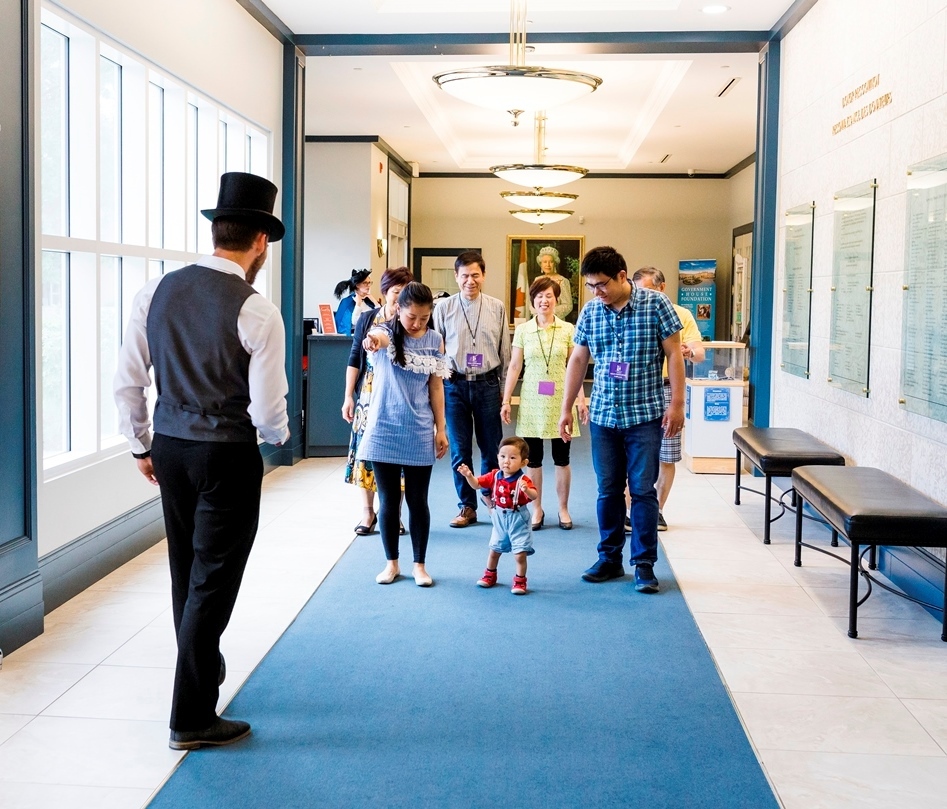 A family group being led by a gentleman in Victorian costume wearing a black top hat on the main level of the QEII wing of Government House SK.